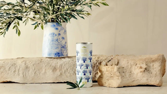 Dekorative Dose mit hochwertigem Olivenöl, verziert mit stilisierten Olivenbäumen, auf einem rustikalen Stein vor einem floralen Keramikgefäß mit frischen Olivenzweigen.