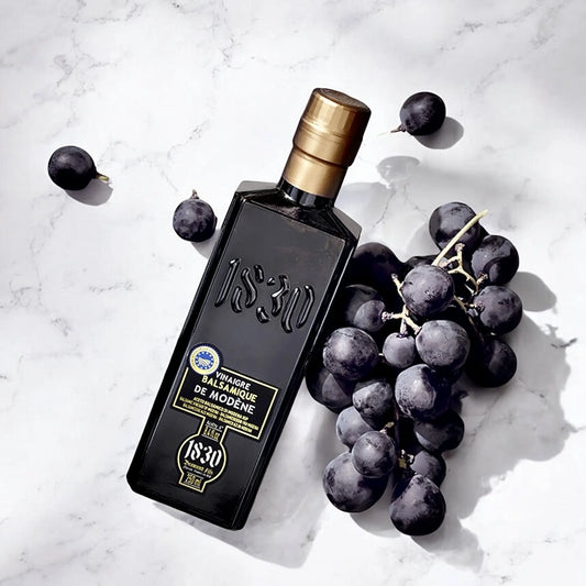 Aceto Balsamico Modena PGI – 3 Jahre gereift, elegant präsentiert auf einem Marmorhintergrund mit frischen dunklen Trauben, die seine hochwertige Herkunft unterstreichen