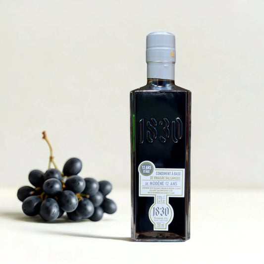 Flasche 12 Jahre gereifter Balsamico Modena neben frischen dunklen Trauben auf hellem Hintergrund. Edler Essig aus Italien mit intensivem Geschmack und cremiger Konsistenz.