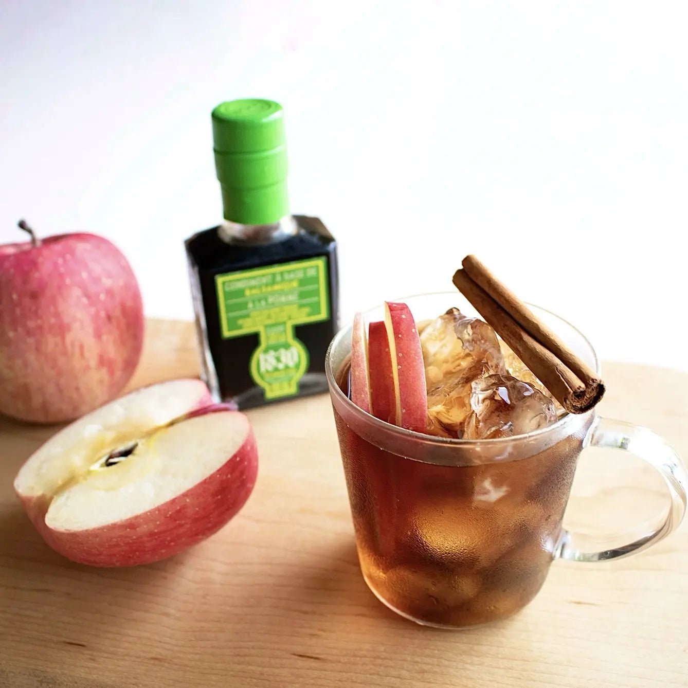 Glas mit Apfelstücken, Eiswürfeln und Zimtstangen neben einer Flasche Apfel Balsamico Essig Condiment 100 ml und einem roten Apfel auf Holzuntergrund. Perfekt für kreative Genussmomente – jetzt Apfel Balsamico Essig kaufen!