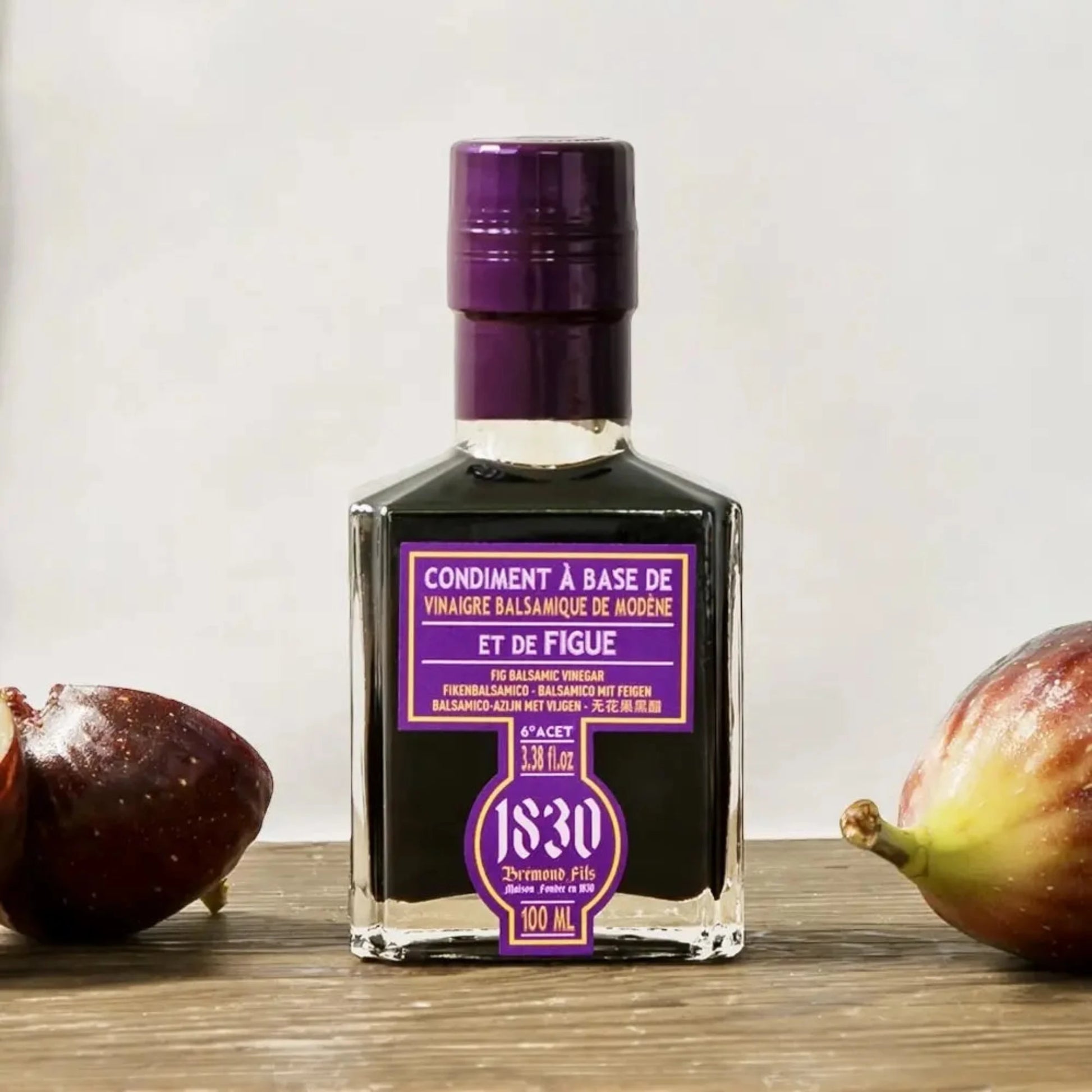 Maison Brémond 1830 Premium Feigen-Balsamico Essig in einer edlen Glasflasche mit lila Etikett, präsentiert mit frischen Feigen auf einem Holztisch.