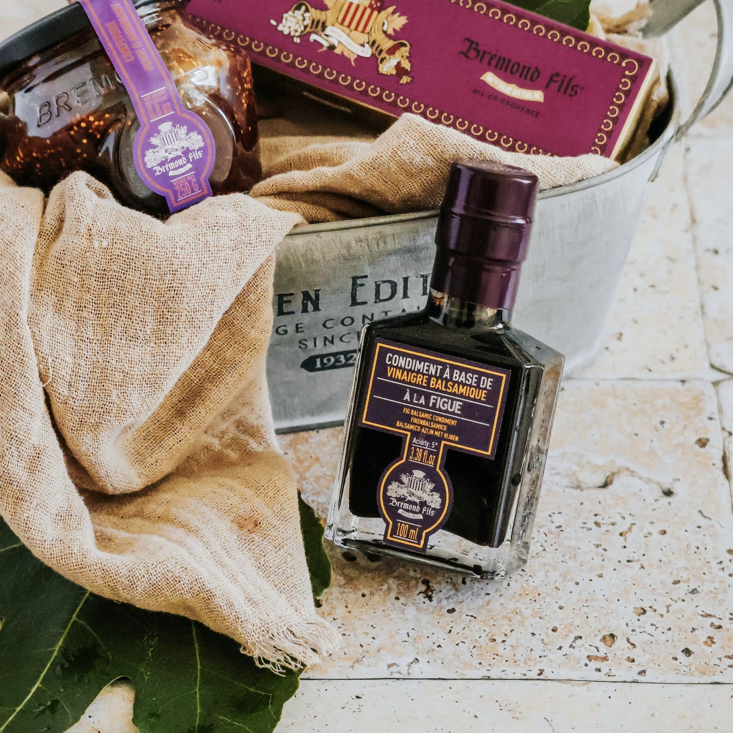 Maison Brémond 1830 Feigen-Balsamico Essig als Teil eines Gourmet-Geschenkkorbs mit anderen Delikatessen, verpackt in rustikalem Leinenstoff.