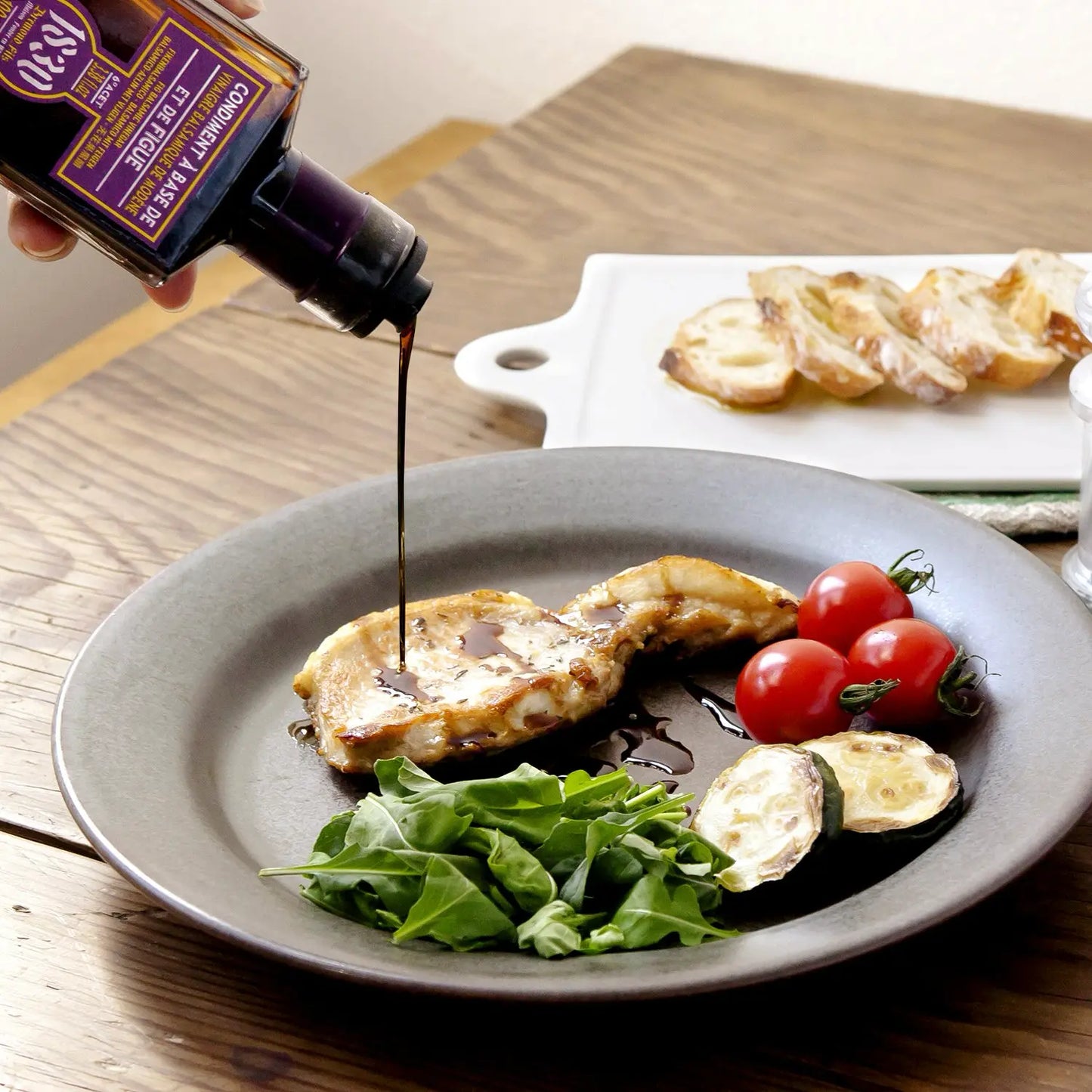 Dunkler Feigen-Balsamico Essig wird aus einer Flasche von Maison Brémond 1830 auf ein Gericht mit gegrilltem Fleisch, Tomaten, Zucchini und Rucola gegossen.