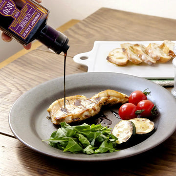 Dunkler Feigen-Balsamico Essig wird aus einer Flasche von Maison Brémond 1830 auf ein Gericht mit gegrilltem Fleisch, Tomaten, Zucchini und Rucola gegossen.