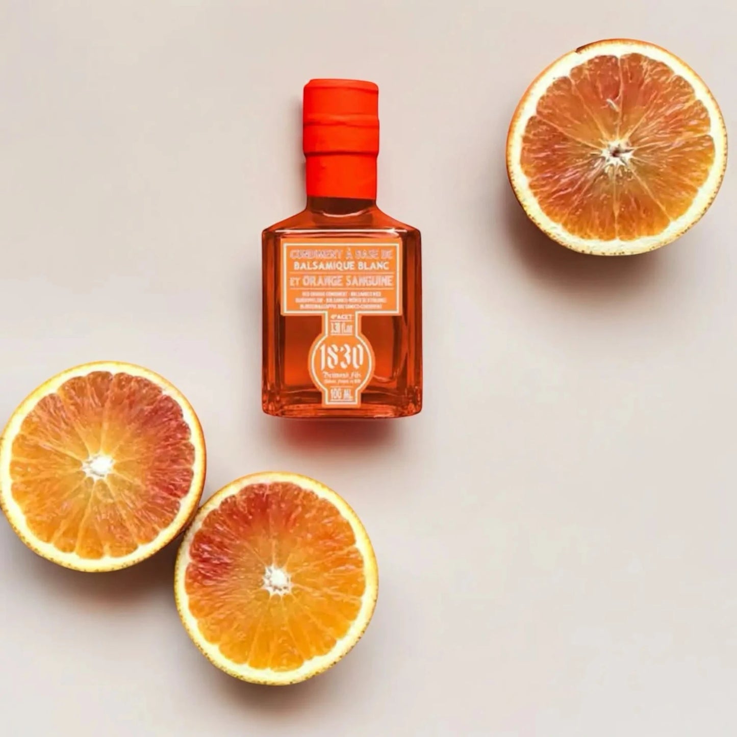 Eine Flasche Blutorange-Balsamico-Essig von Maison Brémond 1830, umgeben von drei aufgeschnittenen Blutorangen auf einem hellen Hintergrund. Die leuchtend orangefarbene Flasche harmoniert mit den frischen Früchten und betont die fruchtige Essenz des Produkts.