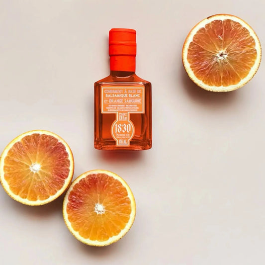 Eine Flasche Blutorange-Balsamico-Essig von Maison Brémond 1830, umgeben von drei aufgeschnittenen Blutorangen auf einem hellen Hintergrund. Die leuchtend orangefarbene Flasche harmoniert mit den frischen Früchten und betont die fruchtige Essenz des Produkts.