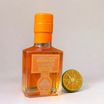 Flasche Kalamansi-Essig neben einer halbierten Kalamansi-Frucht. Die Flasche ist elegant gestaltet, mit orangefarbenem Verschluss und Etikett. Die Kalamansi-Frucht zeigt ihre typische grüne Schale und das orangene Innere.