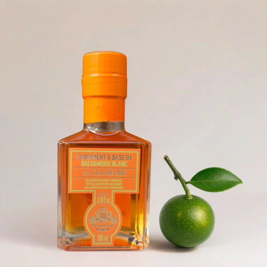 Flasche Kalamansi-Essig neben einer ganzen, frischen Kalamansi-Frucht mit grünem Blatt. Der Essig wirkt hochwertig und exotisch, mit intensivem Orangeton im Flaschenetikett und der Flüssigkeit.