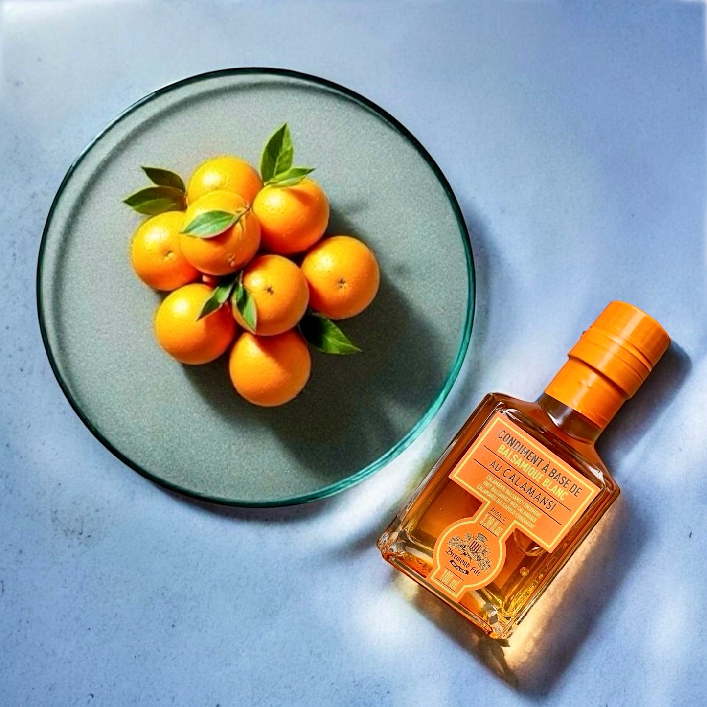 Flasche Kalamansi-Essig neben einer Glasschale mit mehreren frischen, reifen Kalamansi-Früchten. Der Essig hat eine goldgelbe Farbe, das Etikett ist orange und wirkt hochwertig.