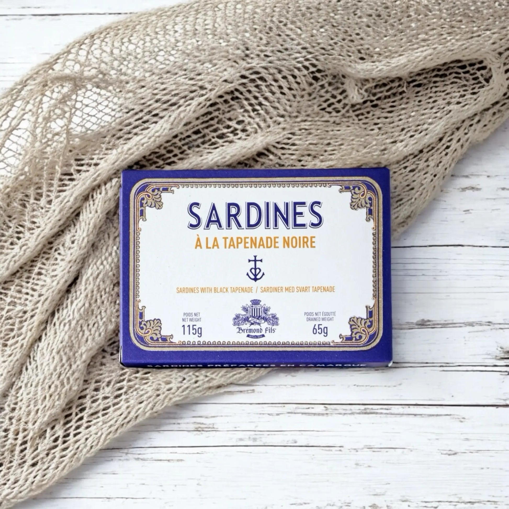 Dose mit französischen Sardinen in schwarzer Tapenade, stilvoll präsentiert auf weißem Holzhintergrund mit maritimem Fischernetz.