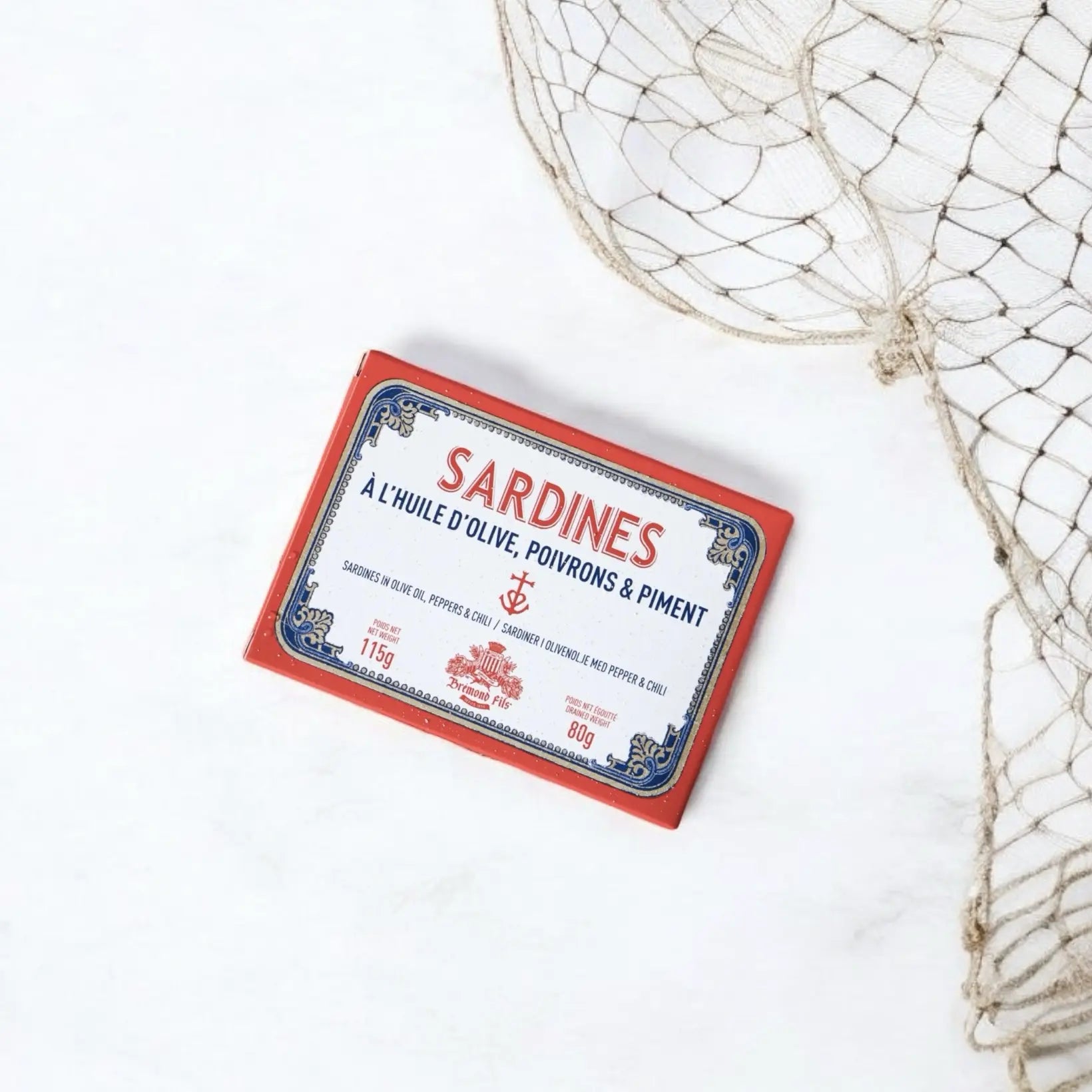 Dose mit Sardinen in nativem Olivenöl, verfeinert mit Paprika und Chili, präsentiert auf einem weißen Untergrund mit Fischernetz-Dekoration. Die Verpackung zeigt ein maritimes Design in Rot, Blau und Weiß mit dem Schriftzug Sardines à l'huile d'olive, poivrons & piment von Maison Brémond 1830.