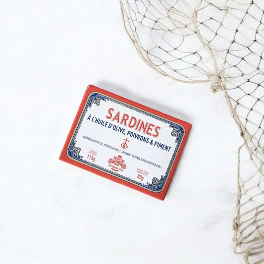 Dose mit Sardinen in nativem Olivenöl, verfeinert mit Paprika und Chili, präsentiert auf einem weißen Untergrund mit Fischernetz-Dekoration. Die Verpackung zeigt ein maritimes Design in Rot, Blau und Weiß mit dem Schriftzug Sardines à l'huile d'olive, poivrons & piment von Maison Brémond 1830.