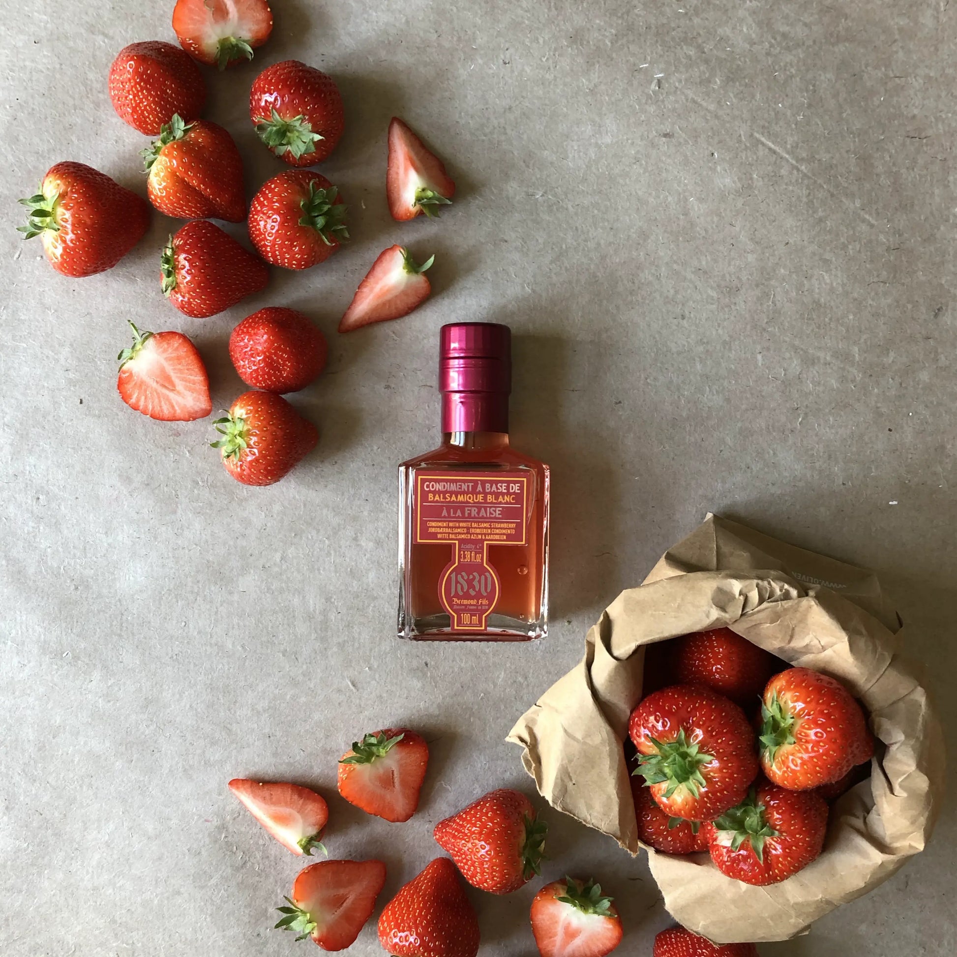 Flasche Erdbeer Balsamico Essig, 100 ml, umgeben von frischen, ganzen und geschnittenen Erdbeeren sowie einem Papiersäckchen mit Erdbeeren. Perfekt für fruchtige und kreative Gerichte.