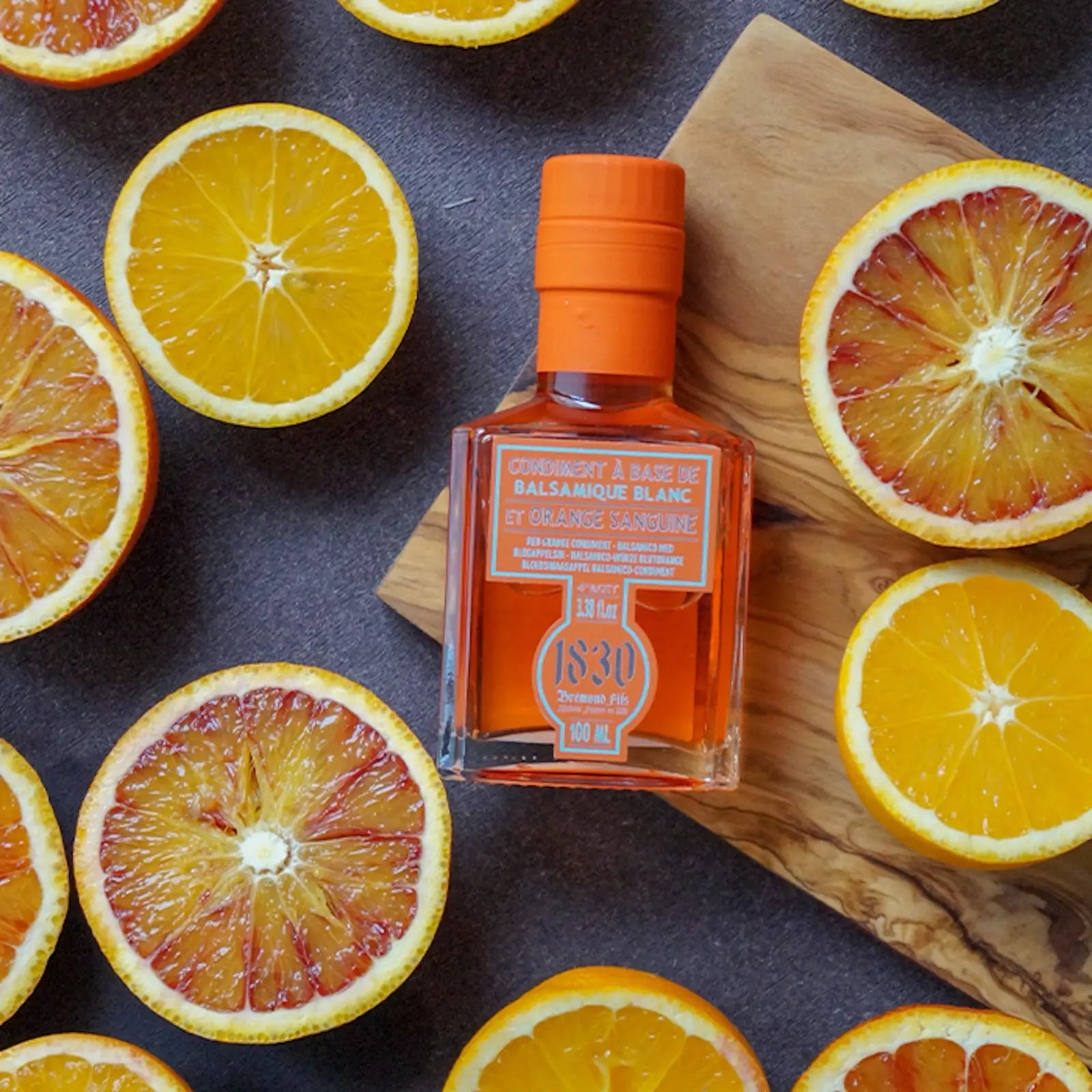 Eine Flasche Blutorange-Balsamico-Essig von Maison Brémond 1830 liegt auf einem Holzbrett, umgeben von frisch aufgeschnittenen Blutorangen. Die leuchtenden Orangetöne des Essigs und der Früchte heben die fruchtige und aromatische Essenz des Produkts hervor.