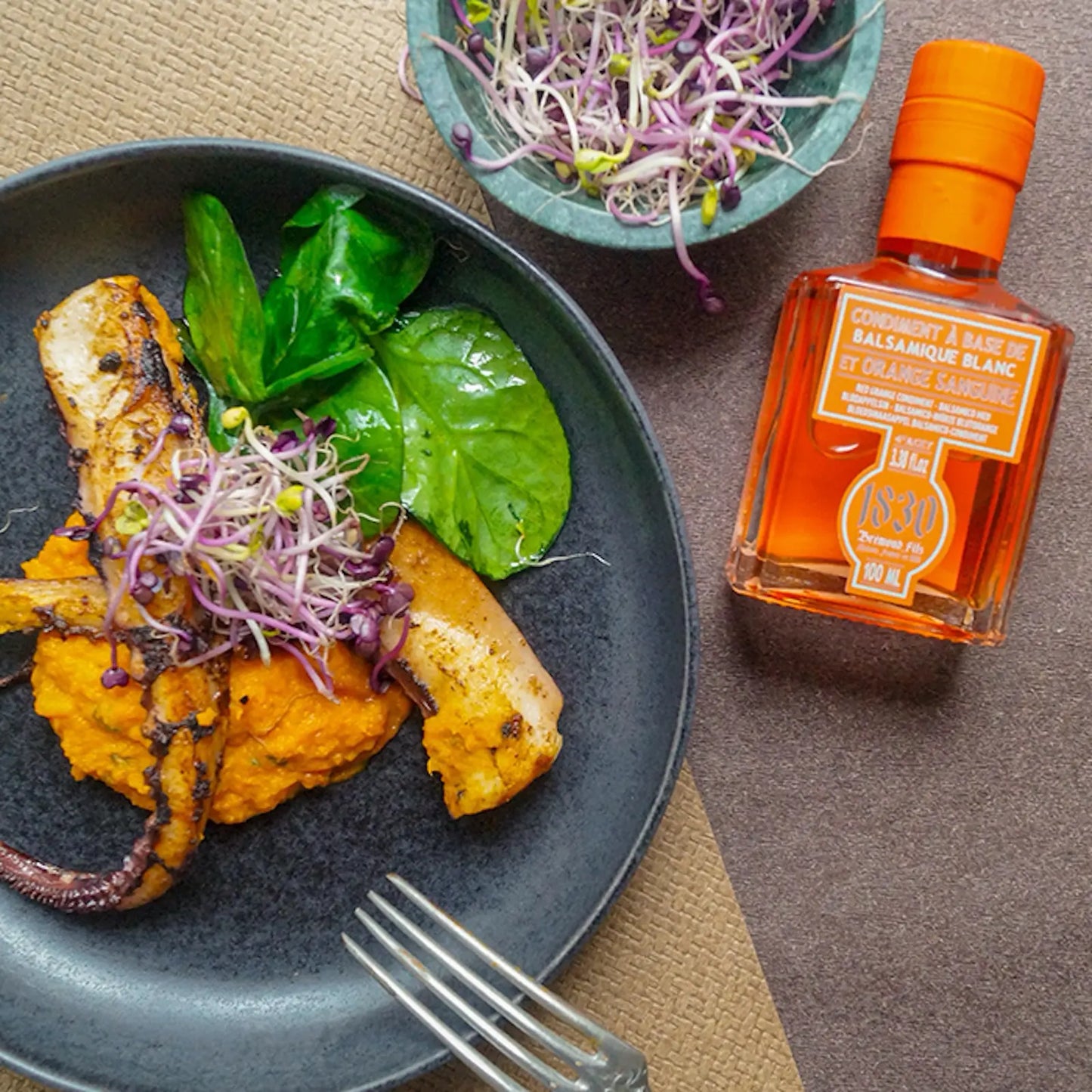 Eine Flasche Blutorange-Balsamico-Essig von Maison Brémond 1830 neben einem stilvoll angerichteten Teller mit gegrilltem Fisch, Kürbispüree, frischem Spinat und Sprossen. Die Szene hebt die vielseitige Verwendung des fruchtigen Balsamico-Essigs in der gehobenen Küche hervor.