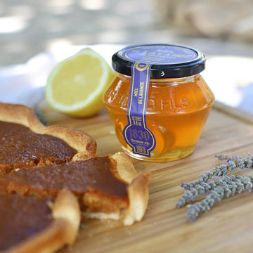 Glas mit Lavendelhonig auf einem Holzbrett, umgeben von einer frisch gebackenen Tarte, einer aufgeschnittenen Zitrone und getrockneten Lavendelblüten. Natürliche, sommerliche Atmosphäre im Freien.