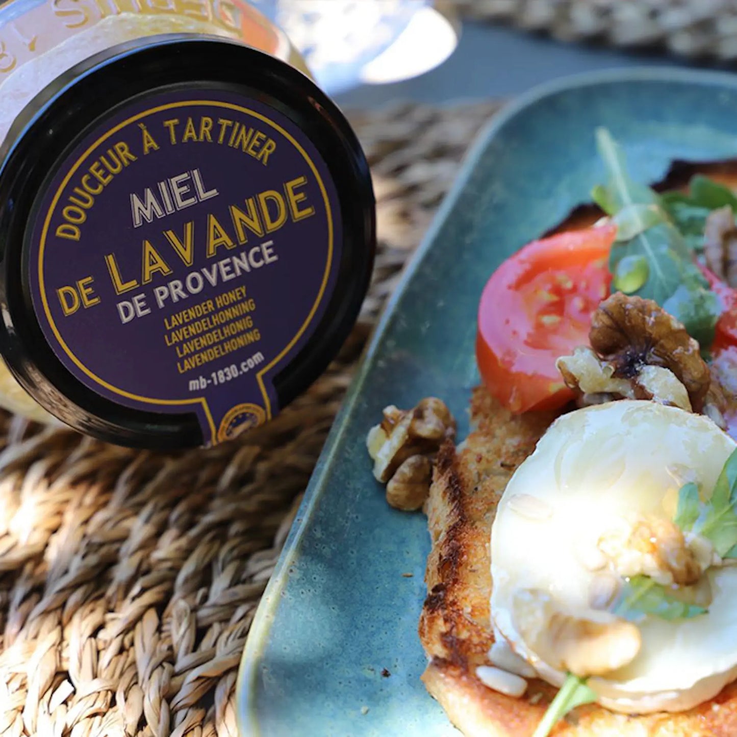 Glas mit Lavendelhonig aus der Provence auf einem geflochtenen Untergrund, daneben eine Scheibe geröstetes Brot mit Ziegenkäse, Tomaten, Walnüssen und Rucola auf einem blauen Teller. Sonnenlicht sorgt für eine warme, mediterrane Atmosphäre.