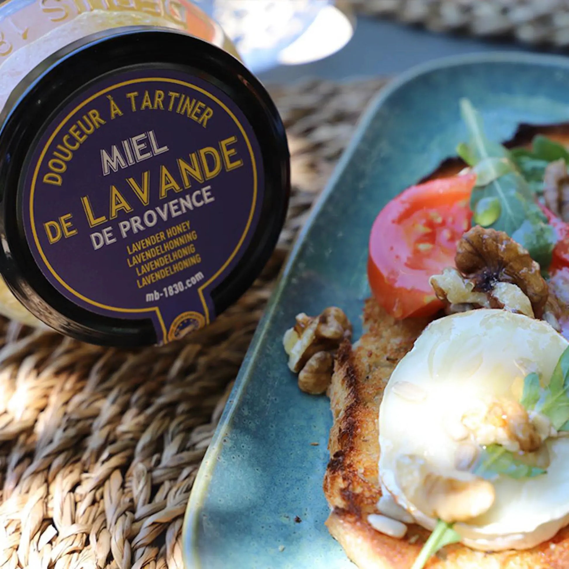 Glas mit Lavendelhonig aus der Provence auf einem geflochtenen Untergrund, daneben eine Scheibe geröstetes Brot mit Ziegenkäse, Tomaten, Walnüssen und Rucola auf einem blauen Teller. Sonnenlicht sorgt für eine warme, mediterrane Atmosphäre.