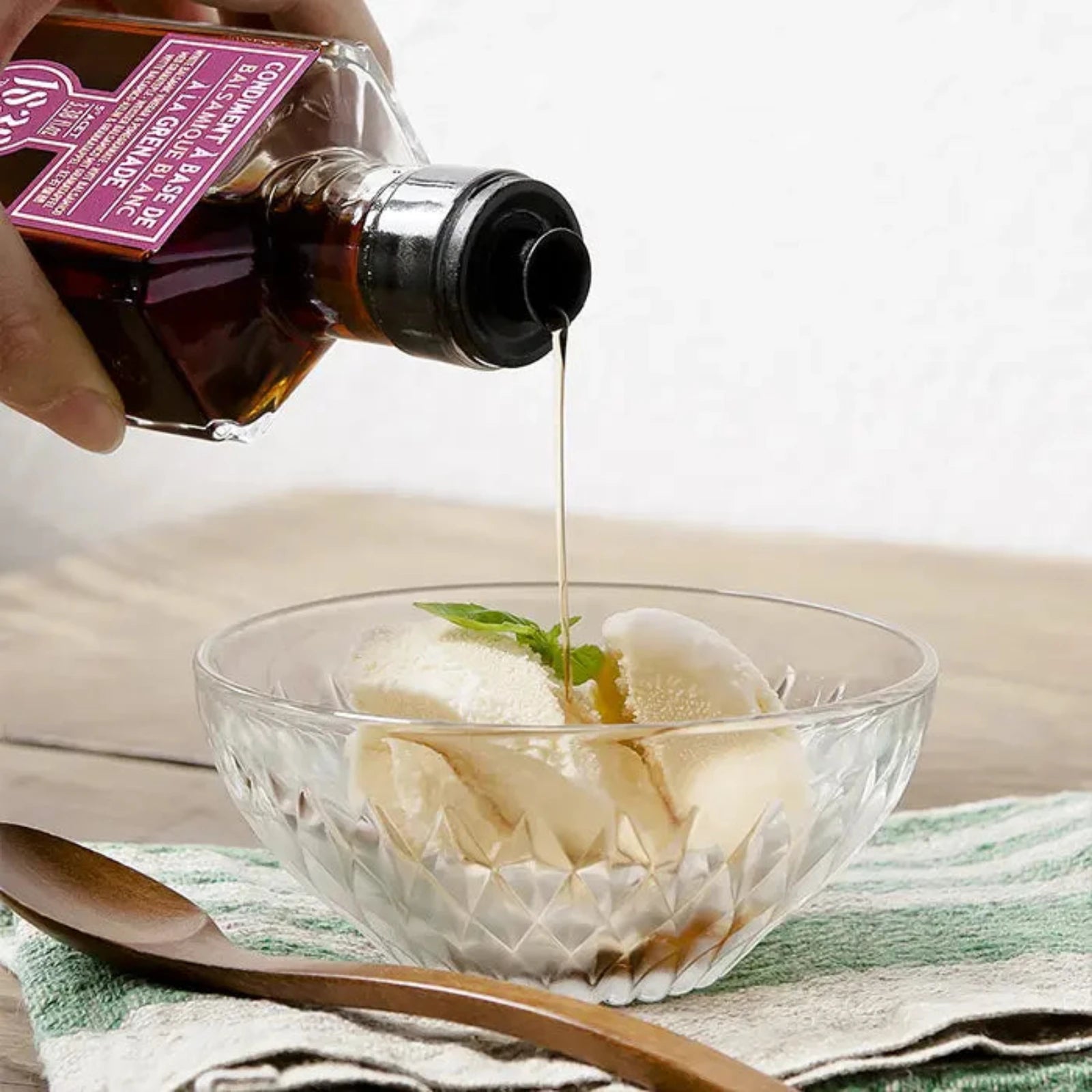 Hand gießt Maison Bremond 1830 Granatapfel-Balsamico-Essig aus einer Flasche auf eine Schale mit Vanilleeis, dekoriert mit Minze.