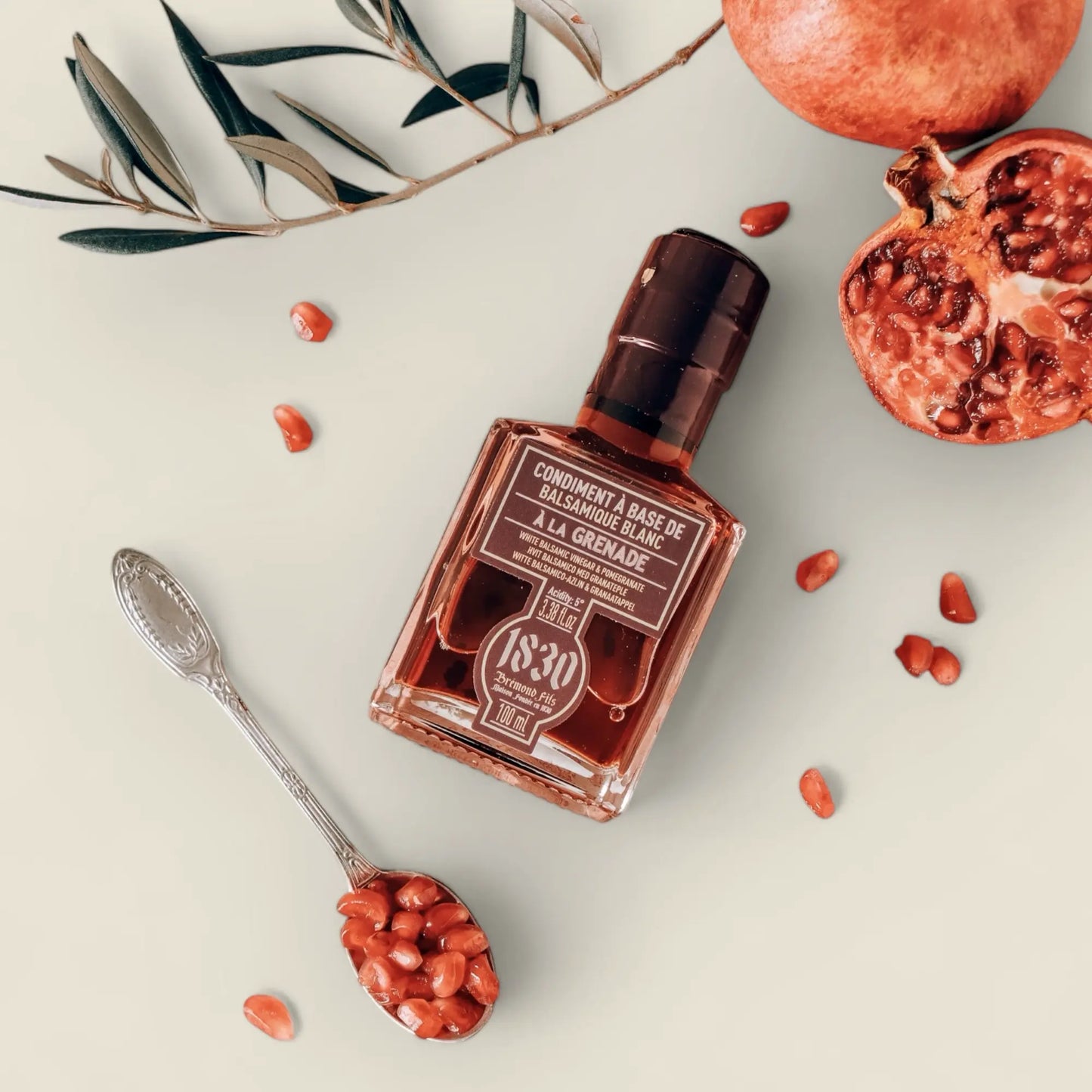 Maison Bremond 1830 Granatapfel-Balsamico-Essig stilvoll präsentiert mit Granatapfel, Olivenzweig und einem silbernen Löffel mit Granatapfelkernen