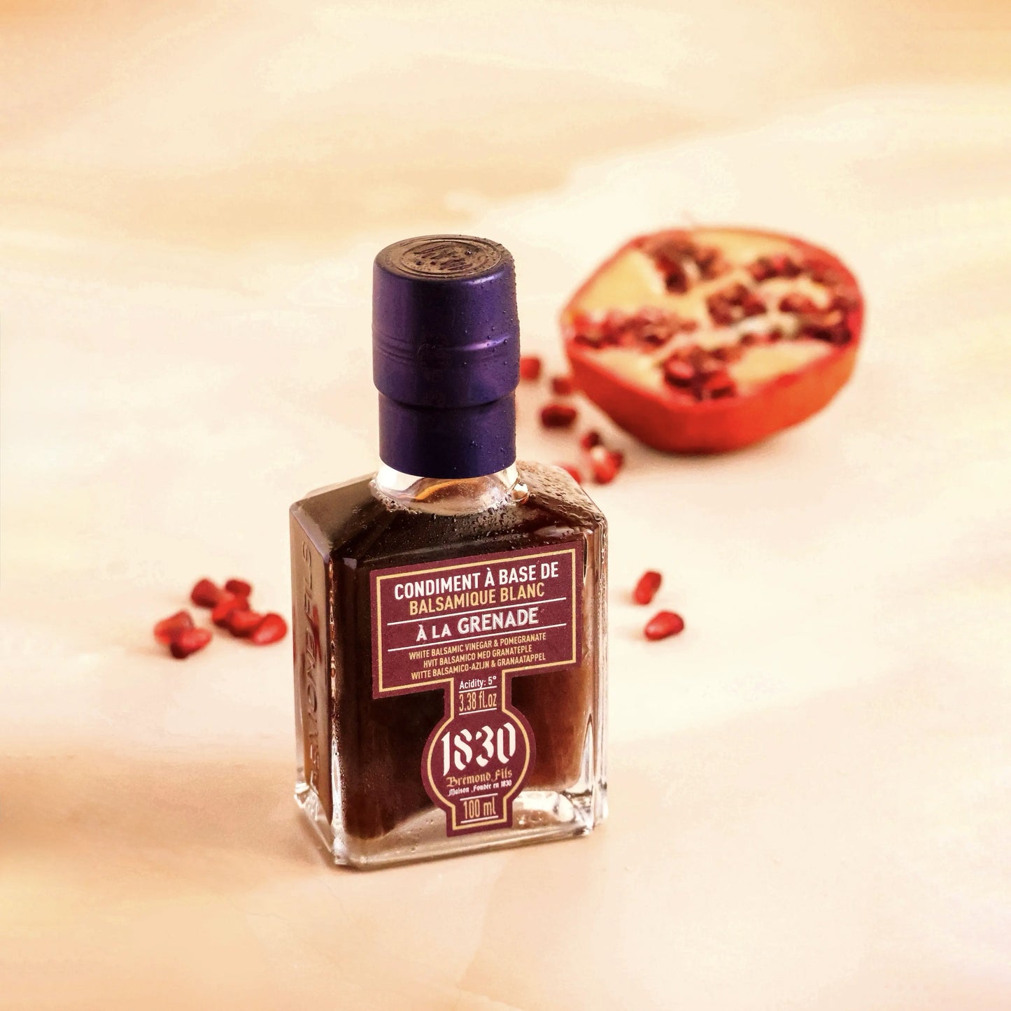 Maison Bremond 1830 Granatapfel-Balsamico-Essig-Flasche vor einem halbierten Granatapfel auf hellem Untergrund, mit verstreuten Granatapfelkernen.