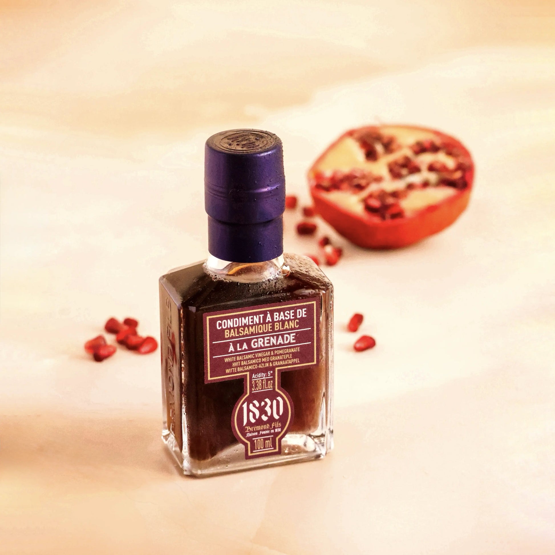 Maison Bremond 1830 Granatapfel-Balsamico-Essig-Flasche vor einem halbierten Granatapfel auf hellem Untergrund, mit verstreuten Granatapfelkernen.