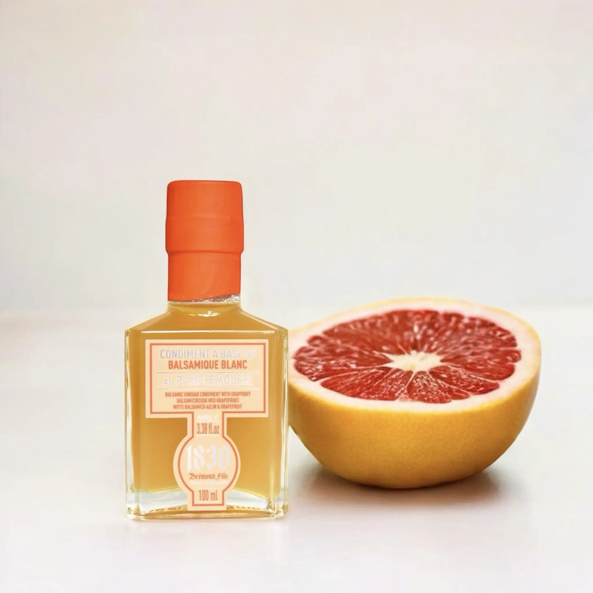 Maison Bremond 1830 Grapefruit-Balsamico-Essig in einer quadratischen Glasflasche, präsentiert mit einer saftigen, frisch aufgeschnittenen Grapefruit auf hellem Hintergrund.