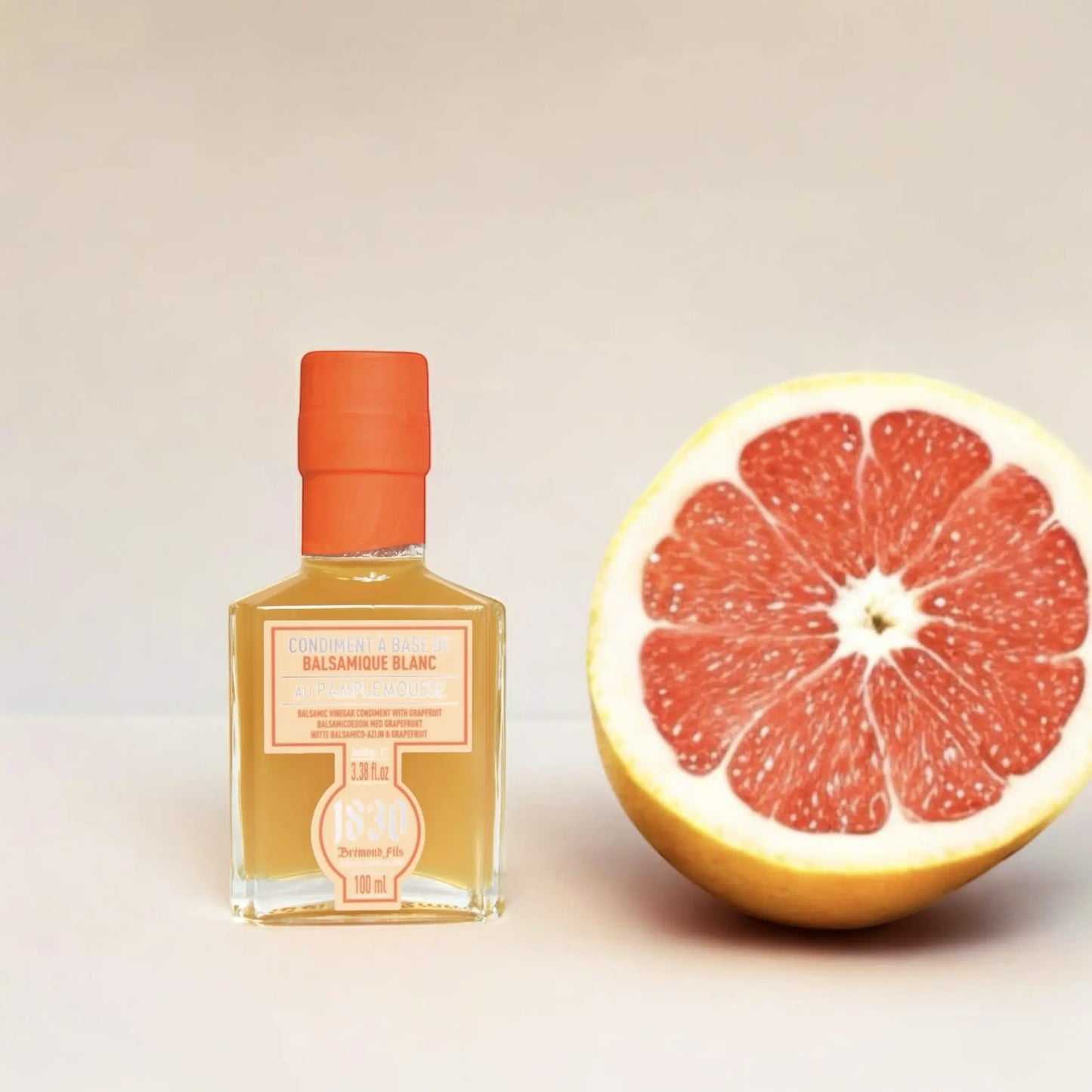 Maison Bremond 1830 Grapefruit-Balsamico-Essig in einer eleganten Glasflasche mit orangefarbenem Etikett und Verschluss, neben einer halbierten roten Grapefruit.