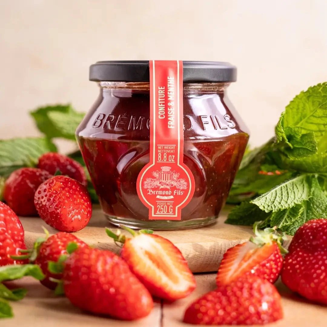 Erdbeer-Minze-Konfitüre im Glas mit rotem Etikett von Maison Brémond, dekoriert mit frischen Erdbeeren und Minzblättern auf einem Holzbrett.