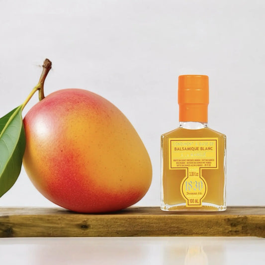 Kleine Glasflasche mit Mango-Balsamico-Essig neben einer frischen, reifen Mango auf einem Holzbrett.
