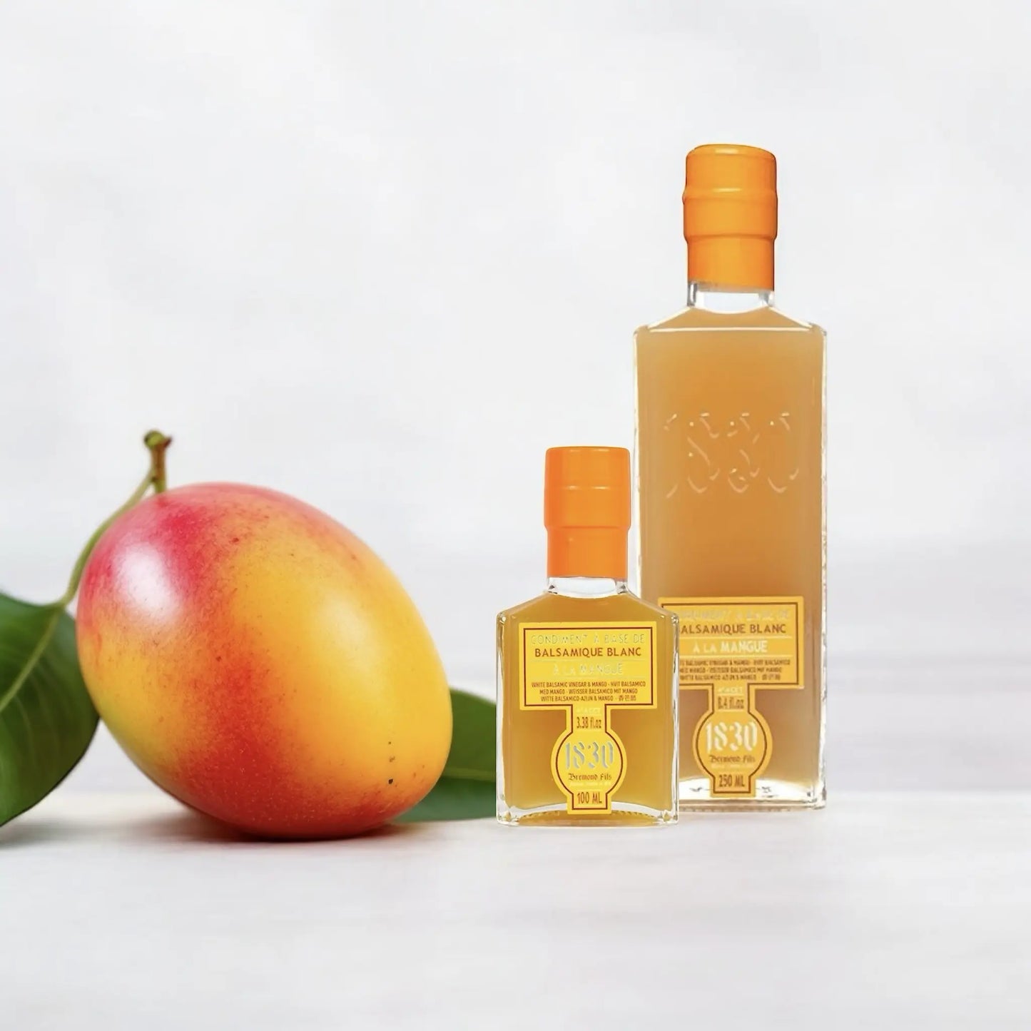 Zwei unterschiedlich große Glasflaschen mit Mango-Balsamico-Essig neben einer frischen Mango, ansprechend arrangiert auf hellem Hintergrund.