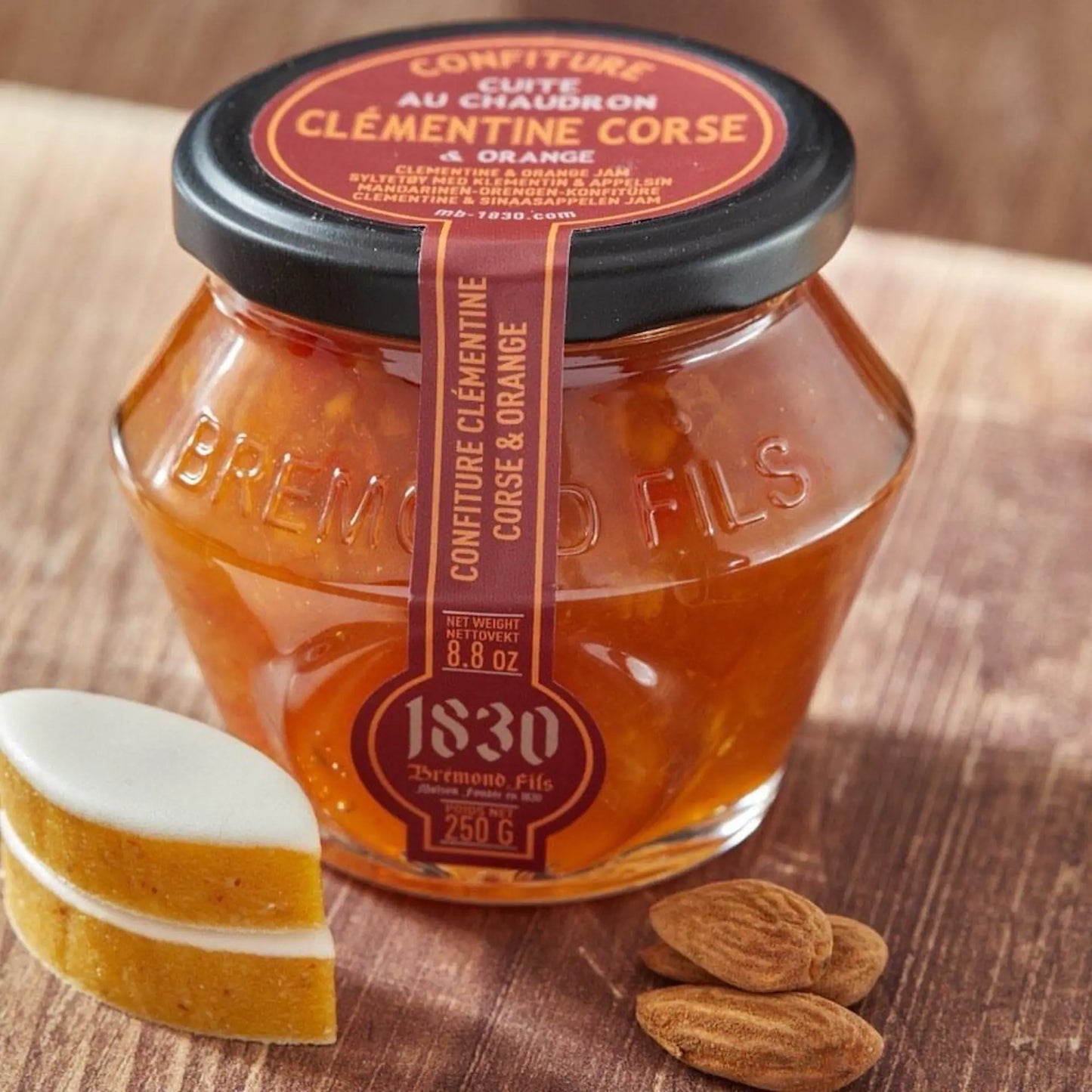 Clementinen-Orangen-Konfitüre auf rustikalem Holztisch, daneben Mandeln und ein aufgeschnittener Calisson. Schwarzer Deckel mit edlem Etikett, leuchtende Marmelade mit Fruchtstücken.