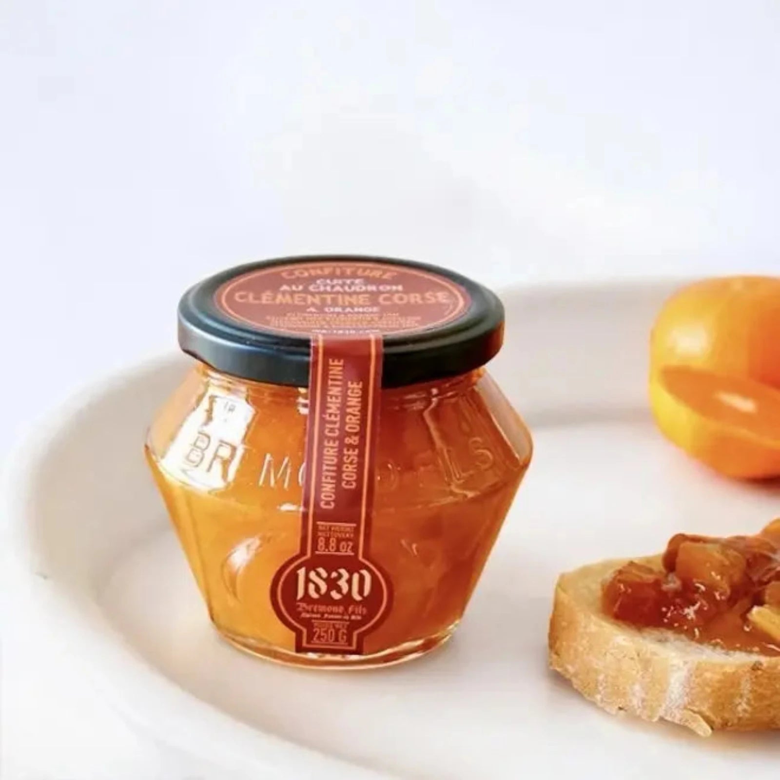 Sechseckiges Glas Clementinen-Orangen-Konfitüre auf weißem Teller, daneben ein Stück Baguette mit Marmelade, im Hintergrund eine aufgeschnittene Clementine.