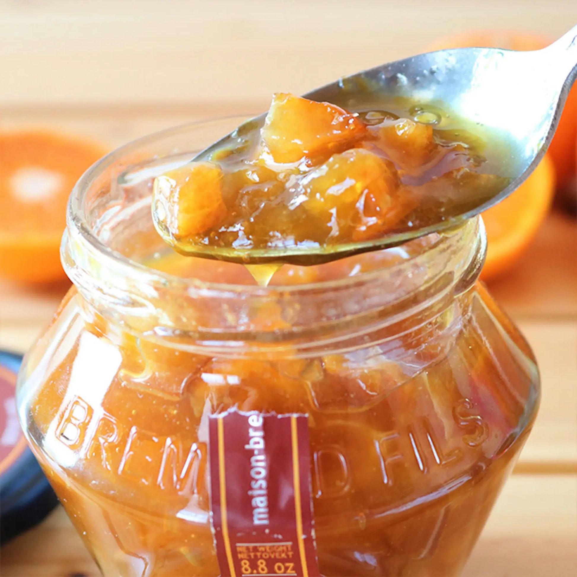 Löffel mit goldgelber Clementinen-Orangen-Marmelade voller Fruchtstücke. Glas mit geprägtem Logo, rote Banderole, im Hintergrund frische Clementinen.