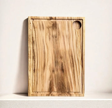 Ein rechteckiges Schneidebrett aus Olivenholz mit einer markanten Maserung, das auf einer hellen Oberfläche liegt. Das Brett hat eine Saftrille entlang der Kanten.