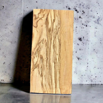 Rechteckiges Schneidebrett aus hochwertigem Olivenholz mit abgerundeten Ecken, aufrecht an eine graue Betonwand gelehnt, mit einer glänzenden Oberfläche im Vordergrund. Die natürliche Maserung des Holzes ist deutlich sichtbar.