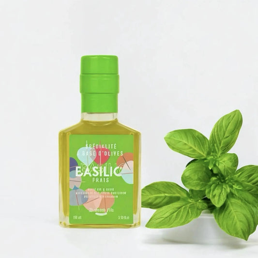 Kleine Flasche Olivenöl Basilikum (100 ml) mit grünem Deckel, daneben frisches Basilikum in einem weißen Topf. Frisches, aromatisiertes Olivenöl für mediterrane Gerichte.