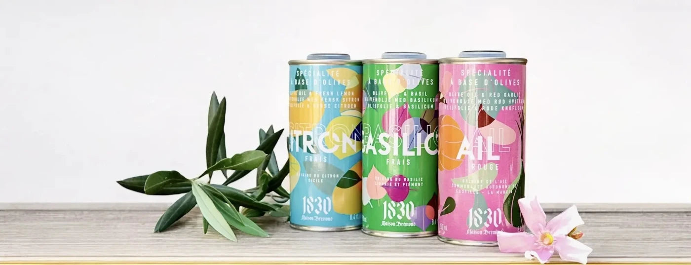 Drei farbenfrohe Dosen mit aromatisiertem Olivenöl kaufen der Marke Maison Brémond 1830. Die Geschmacksrichtungen umfassen Olivenöl Zitrone, Olivenöl Basilikum und Olivenöl Knoblauch, stilvoll präsentiert mit einem Olivenzweig und einer rosa Blüte auf hellem Hintergrund. Ideal für Feinschmecker und mediterrane Küche.