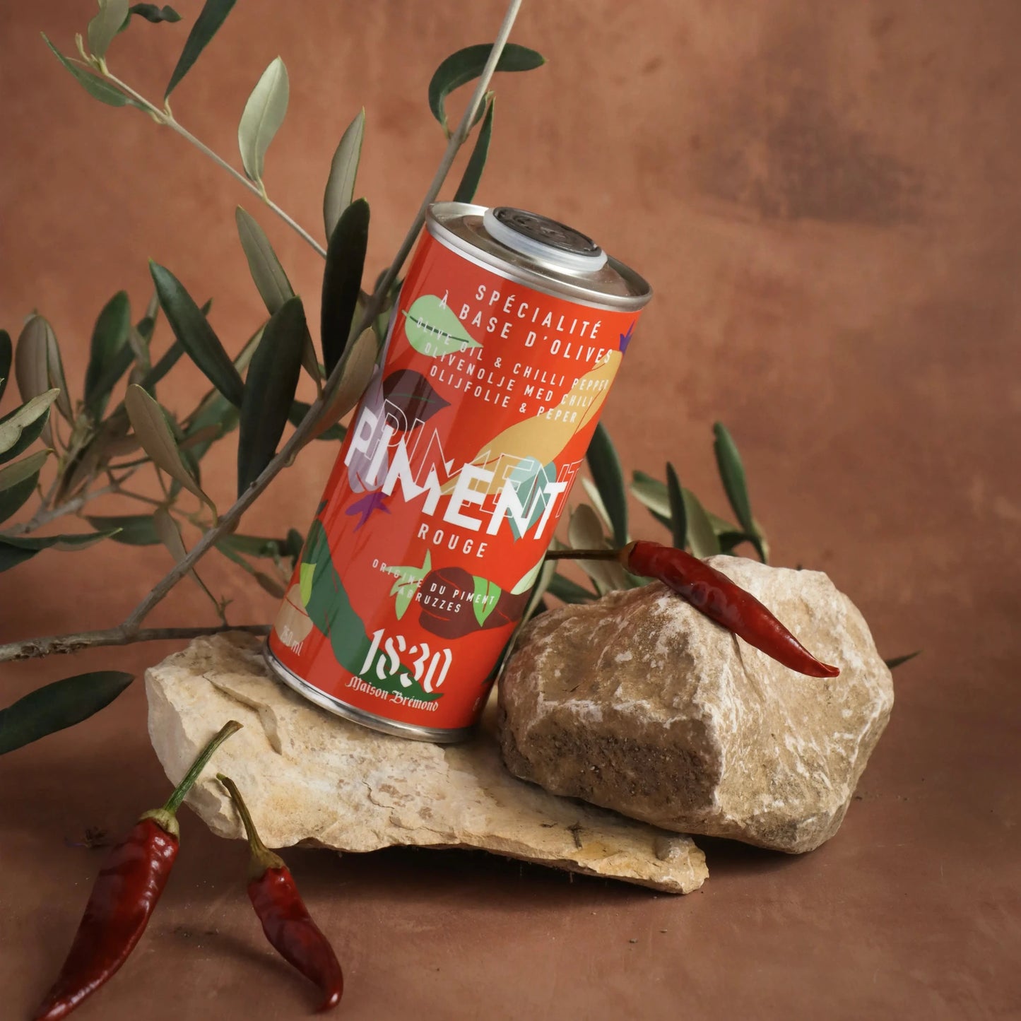 Maison Brémond Olivenöl mit Chili 250 ml – Feuriges Chili-Olivenöl in einem stilvollen roten Kanister, inszeniert mit Olivenzweigen und scharfen Chilischoten auf Natursteinen.
