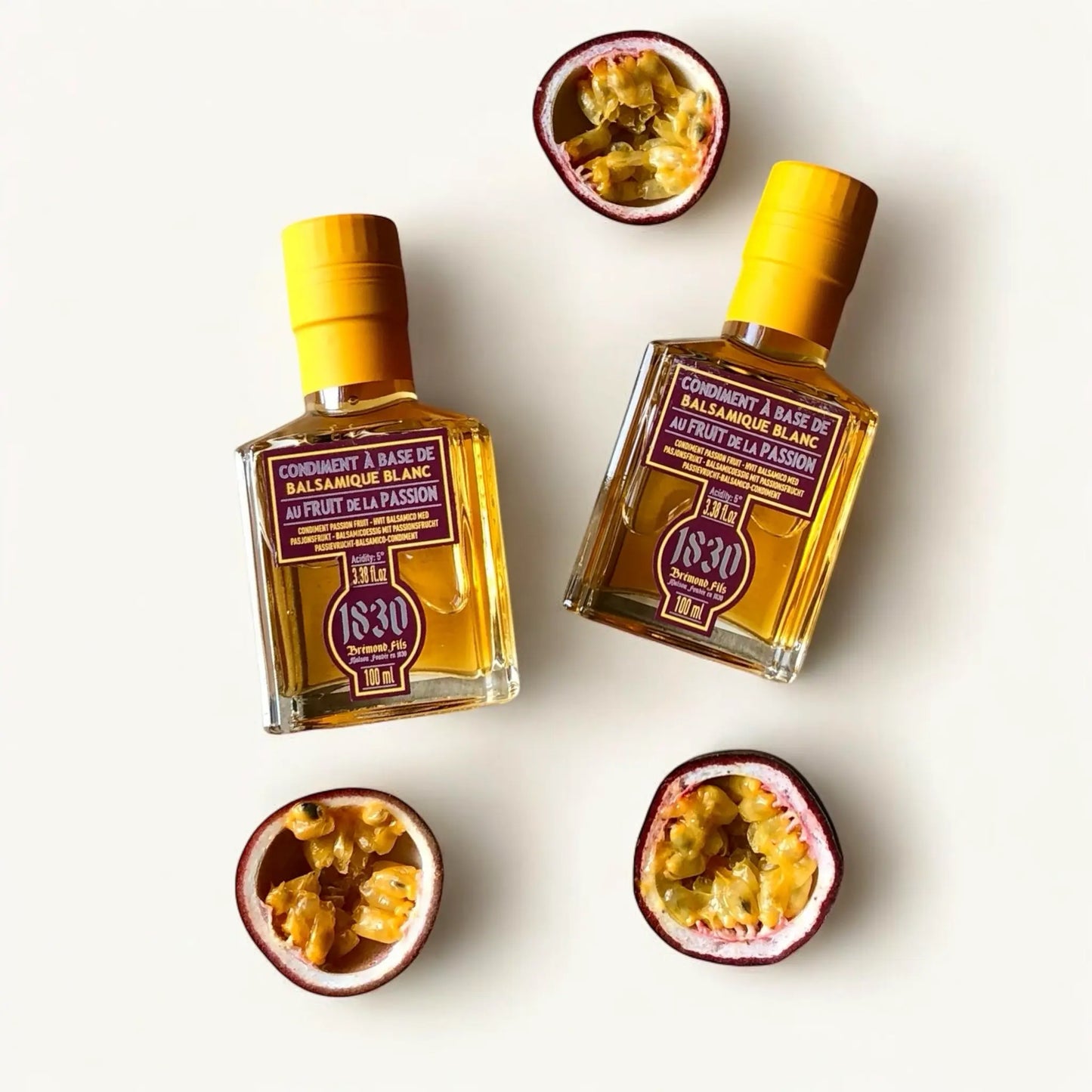 Zwei stilvolle Flaschen weißer Balsamico-Essig Condiment mit Passionsfrucht von Maison Brémond 1830, dekoriert mit frischen, aufgeschnittenen Passionsfrüchten auf hellem Hintergrund. Perfekt für Feinschmecker und Liebhaber tropischer Aromen.