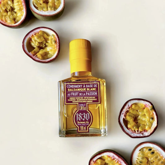 Flasche weißer Balsamico-Essig Condiment mit Passionsfrucht von Maison Brémond 1830, dekoriert mit aufgeschnittenen, frischen Passionsfrüchten auf hellem Hintergrund. Tropischer Genuss für Feinschmecker und Gourmets.