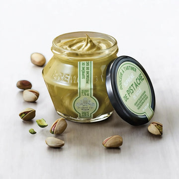 Offenes Glas Pistaziencreme mit cremiger Konsistenz, umgeben von ganzen Pistazien auf einer rustikalen Holzoberfläche.