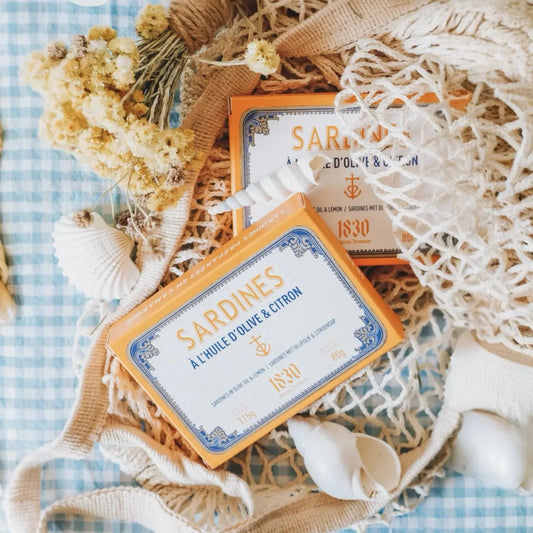 Dose Sardinen in Olivenöl & Zitrone, stilvoll arrangiert mit maritimen Elementen wie Muscheln und Netzen auf kariertem Stoff. Hochwertige Verpackung mit klassischem Design.