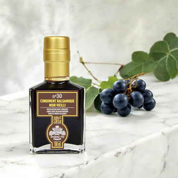 Luxuriöser schwarzer Balsamico-Essig – 30 Jahre gereift, präsentiert auf einer weißen Marmorplatte mit frischen Trauben.
