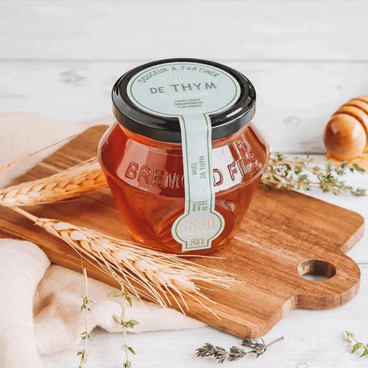 Ein Glas mit 250 g Thymianhonig, präsentiert von Maison Brémond 1830. Der Honig hat eine klare, hell-bernsteinfarbene Erscheinung und wird in einem transparenten Glasgefäß mit einem goldfarbenen Deckel angeboten. Auf dem Etikett sind der Produktname und das Logo des Herstellers zu sehen. Der Honig stammt aus Katalonien, Spanien, und zeichnet sich durch kräftige, krautige Noten aus