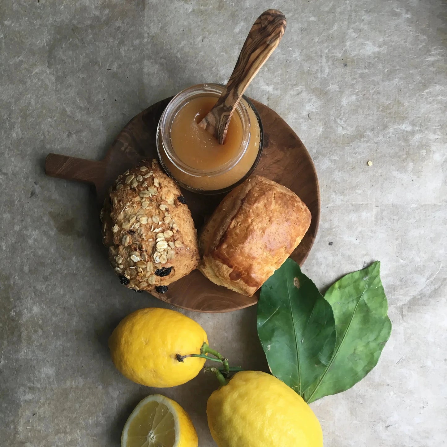 Maison Brémond Zitronencreme (Lemon Curd) in einem offenen Glas mit Holzlöffel, serviert mit frischen Brötchen und Zitronen – cremig, fruchtig und perfekt für Frühstück & Gebäck.