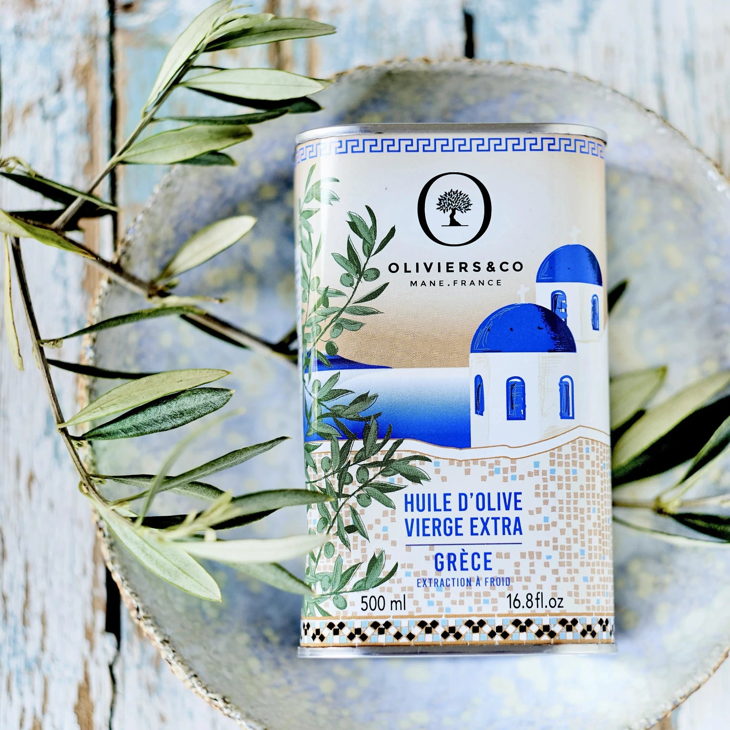 500 ml Dose griechisches Olivenöl von Oliviers & Co, dekoriert mit einem Bild einer traditionellen blau-weißen Kapelle und umgeben von Olivenzweigen. Die Dose liegt auf einem rustikalen Teller, was die Authentizität und hohe Qualität des extra nativen Olivenöls betont. Ideal für mediterrane Gerichte und Liebhaber hochwertiger, sortenreiner Olivenöle aus Griechenland.