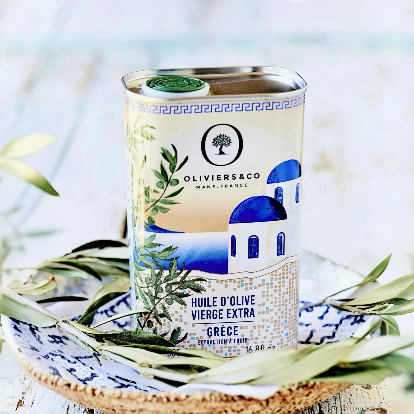 500 ml Dose griechisches Olivenöl von Oliviers & Co, stilvoll präsentiert auf einem rustikalen Teller mit Olivenzweigen. Die Verpackung zeigt eine traditionelle blau-weiße Kapelle und betont die hohe Qualität des extra nativen Olivenöls aus Griechenland. Ideal zum Kaufen für authentische mediterrane Küche.