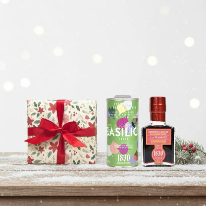 Geschenk mit roter Schleife, Basilikum-Olivenöl und Himbeer-Balsam-Essig von Maison Brémond 1830, festlich arrangiert auf verschneitem Holztisch vor winterlichem Hintergrund.