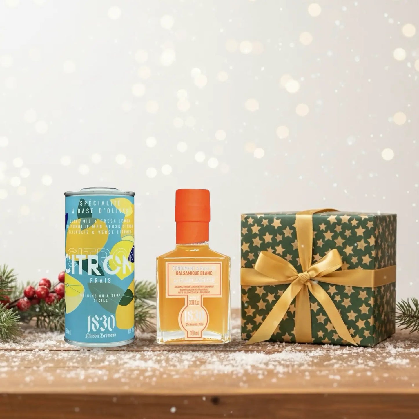 Geschenk mit goldener Schleife, Olivenöl mit Zitrone und Grapefruit-Balsam-Essig von Maison Brémond 1830, festlich dekoriert auf verschneitem Holztisch mit Tannenzweig und roten Beeren im Hintergrund.
